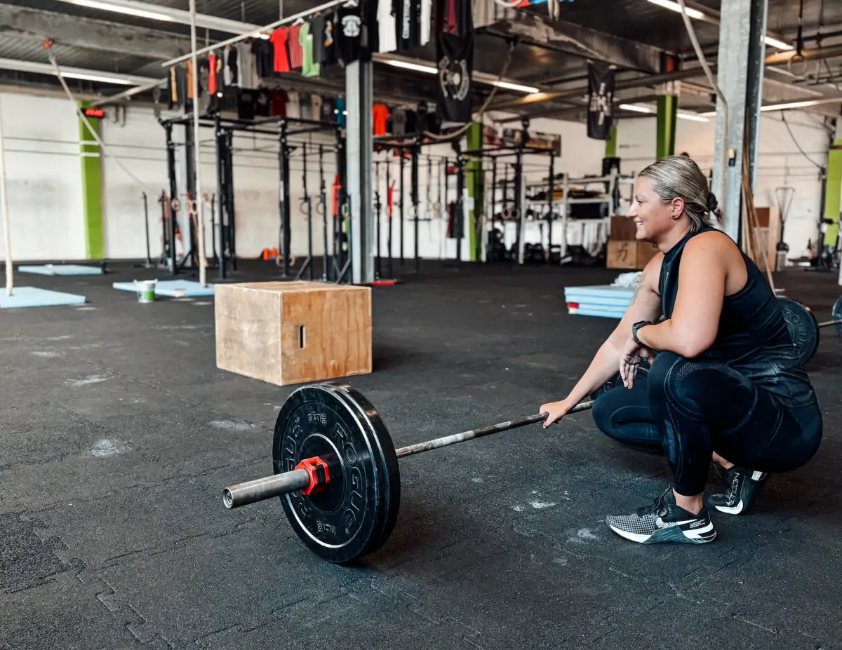 Smiling athlete crouching next to a loaded barbell in a large CrossFit box
