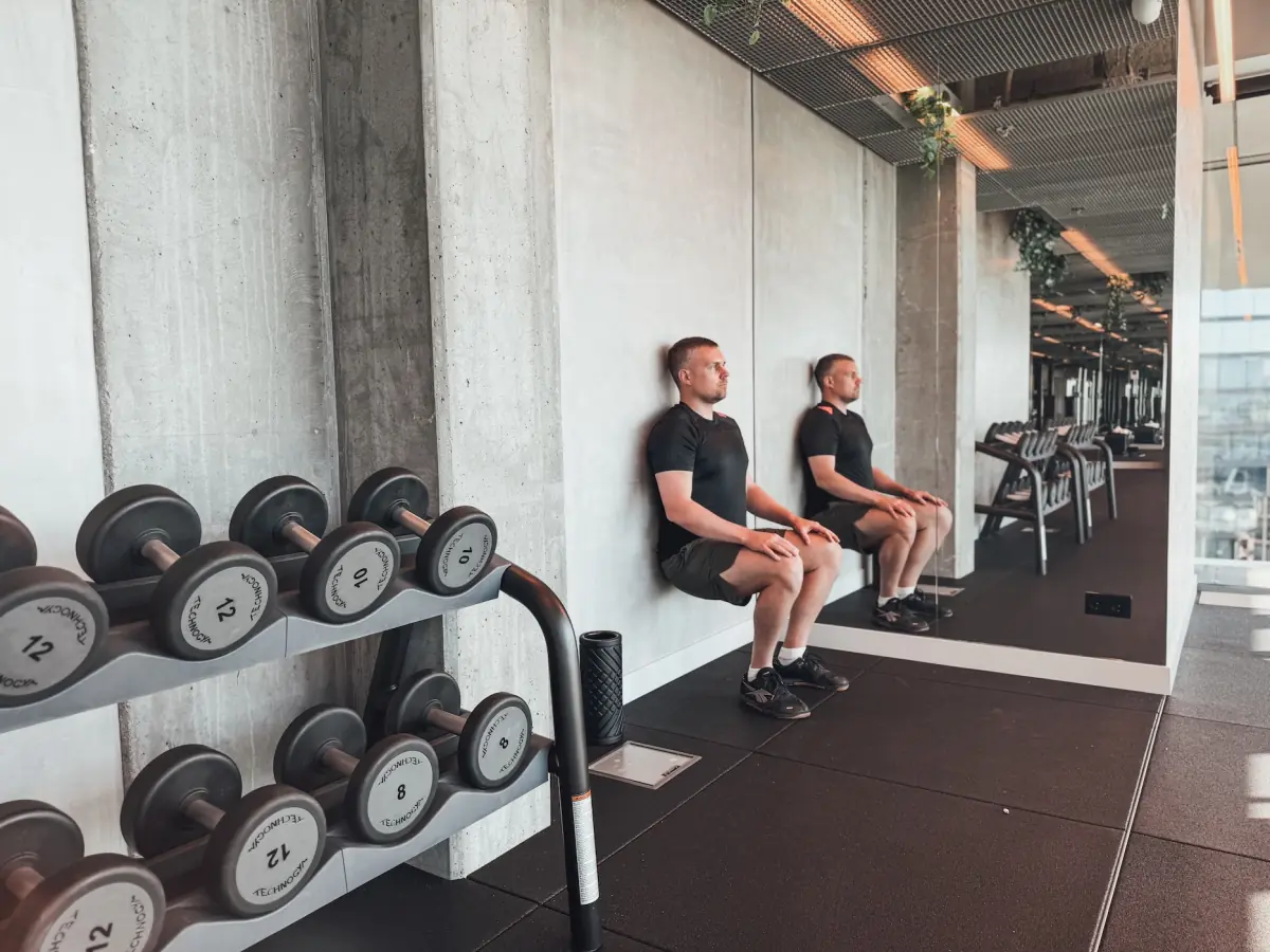 Athlete holding a wall sit against a concrete wall next to a dumbbell rack in the gym