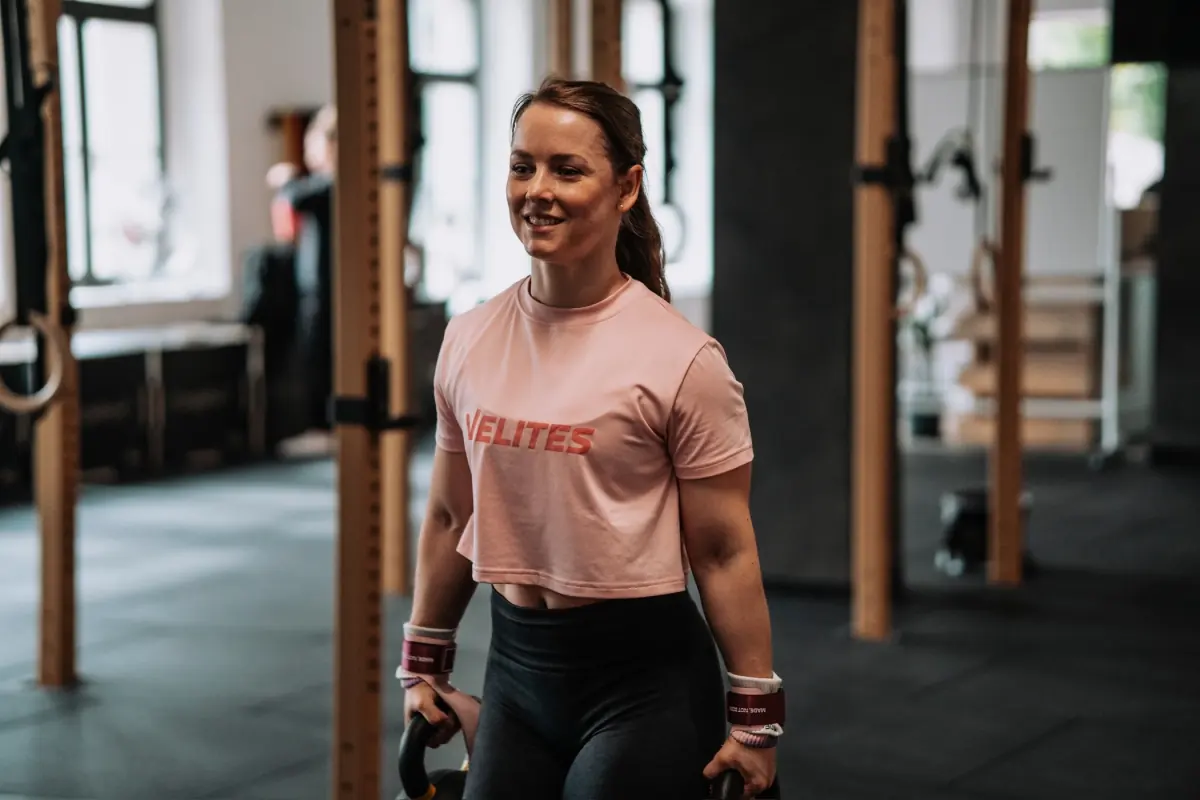 Lächelnde Athletin trägt beim Farmer’s Carry zwei Kettlebells durch das Gym